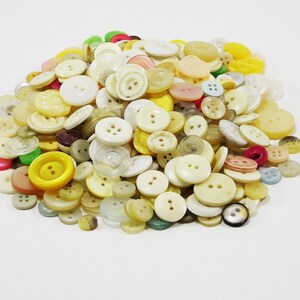 Può includere: Un mucchio di bottoni assortiti in varie dimensioni e tonalità di giallo, crema e bianco sporco. Alcuni bottoni hanno due fori, altri quattro, e alcuni hanno un design a spirale. Alcuni bottoni sono rosa, verdi e blu.