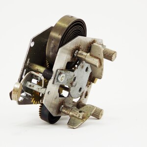 May include: Close-up of a silver and brass clockwork mechanism with a coiled spring and gears. The mechanism is disassembled and shows the inner workings of the clock.