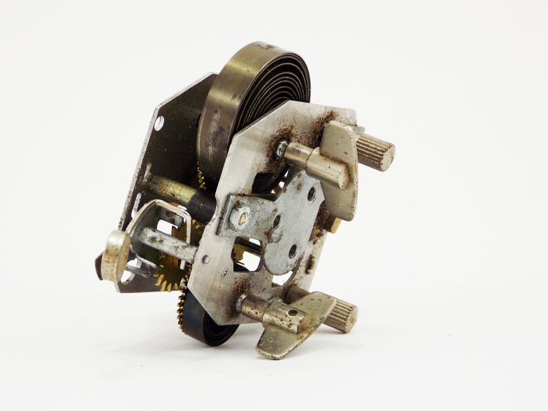 May include: Close-up of a silver and brass clockwork mechanism with a coiled spring and gears. The mechanism is disassembled and shows the inner workings of the clock.