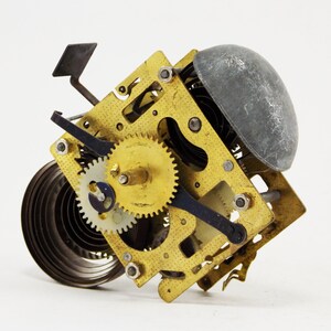 Könnte beinhalten: Nahaufnahme eines goldfarbenen Uhrwerks mit einer Feder, Zahnrädern und einer silberfarbenen Glocke. Der Mechanismus ist zerlegt und die Teile sind sichtbar.