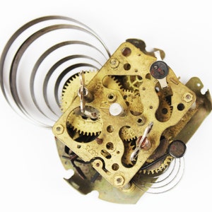 May include: Close-up of a disassembled clock mechanism with brass gears and a coiled spring. The mechanism is on a white background.