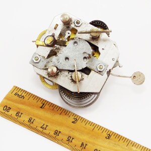 May include: Close-up of a silver and gold clockwork mechanism with a spring and gears. The mechanism is about 2.5 cm in diameter and is sitting on a wooden ruler.