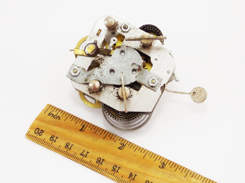 May include: Close-up of a silver and gold clockwork mechanism with a spring and gears. The mechanism is about 2.5 cm in diameter and is sitting on a wooden ruler.