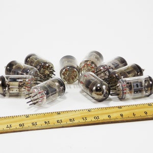 May include: A group of nine vintage vacuum tubes, some with glass casings and some with metal casings, are scattered on a white surface next to a wooden ruler.