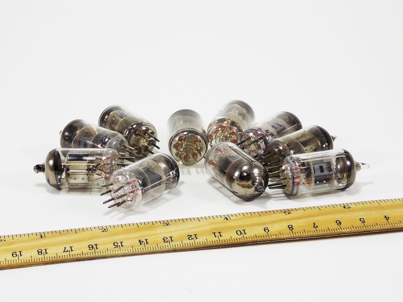 May include: A group of nine vintage vacuum tubes, some with glass casings and some with metal casings, are scattered on a white surface next to a wooden ruler.