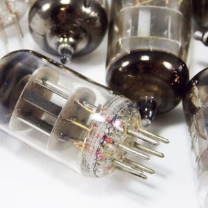May include: Close-up of a group of vintage vacuum tubes, with a focus on one tube with a clear glass body and multiple pins. The tube has a dark grey metal base and a clear glass top with visible internal components.