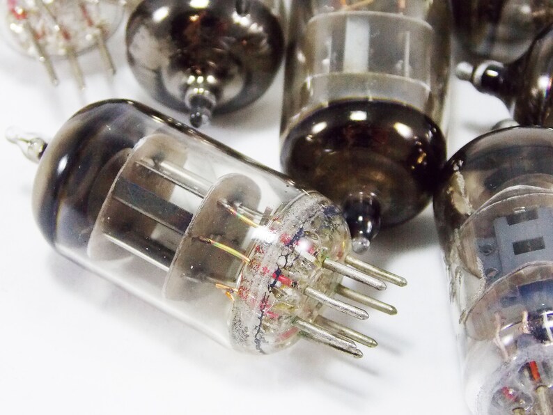 May include: Close-up of a group of vintage vacuum tubes, with a focus on one tube with a clear glass body and multiple pins. The tube has a dark grey metal base and a clear glass top with visible internal components.