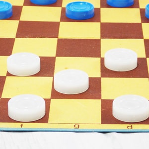 Pode incluir: Um jogo de damas em andamento com pe&ccedil;as brancas e azuis. O tabuleiro tem um padr&atilde;o quadriculado marrom e amarelo. As letras 'f', 'g' e 'd' s&atilde;o vis&iacute;veis no tabuleiro.