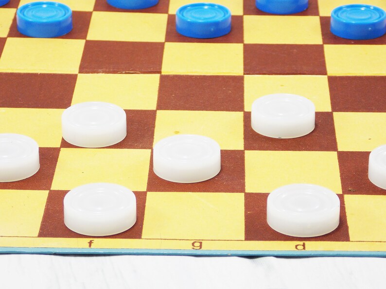Pode incluir: Um jogo de damas em andamento com pe&ccedil;as brancas e azuis. O tabuleiro tem um padr&atilde;o quadriculado marrom e amarelo. As letras 'f', 'g' e 'd' s&atilde;o vis&iacute;veis no tabuleiro.