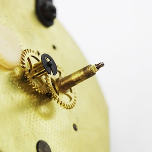 May include: Close-up of a gold-coloured clock mechanism with a small, black gear and a brass rod with a pointed end.