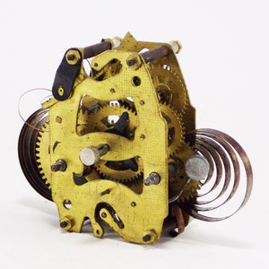 May include: Close-up of a gold-coloured clock mechanism with a spring and gears. The mechanism is made of metal and has a worn, antique look.