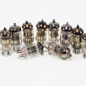 May include: A group of nine vintage vacuum tubes with glass enclosures and metal caps. The tubes have a silver finish and are arranged in a semi-circle on a white surface.