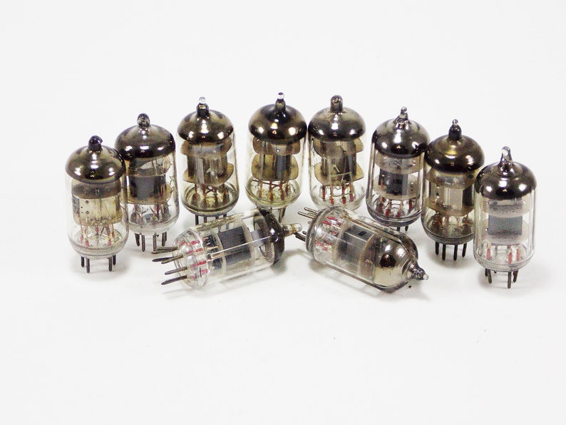 May include: A group of nine vintage vacuum tubes with glass enclosures and metal caps. The tubes have a silver finish and are arranged in a semi-circle on a white surface.