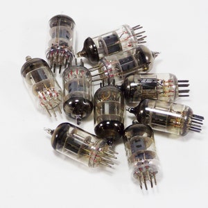 May include: A group of ten vintage vacuum tubes with glass casings and metal pins. The tubes are arranged in a pile on a white surface.
