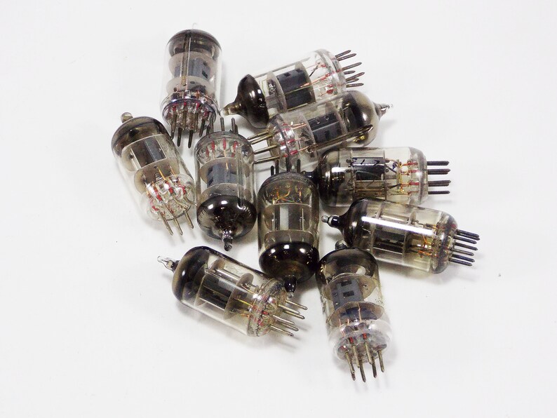 May include: A group of ten vintage vacuum tubes with glass casings and metal pins. The tubes are arranged in a pile on a white surface.