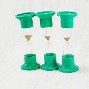 May include: Three green hourglass timers with clear glass bulbs and a brown sand inside. The hourglasses are on a white surface.