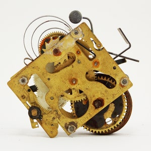 May include: Close-up of a gold-toned clock mechanism with intricate gears and a spring. The mechanism is disassembled and shows the inner workings of the clock.