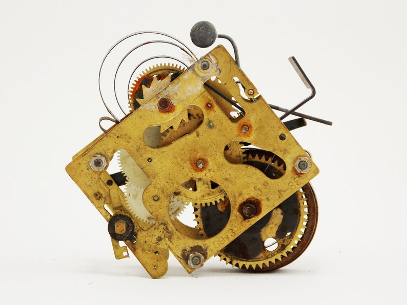 May include: Close-up of a gold-toned clock mechanism with intricate gears and a spring. The mechanism is disassembled and shows the inner workings of the clock.