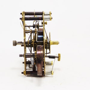 May include: A close-up of a brass clockwork mechanism with gears, springs, and a winding key. The mechanism is disassembled and laid out on a white surface.