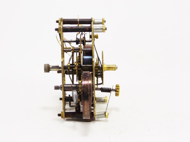 May include: A close-up of a brass clockwork mechanism with gears, springs, and a winding key. The mechanism is disassembled and laid out on a white surface.
