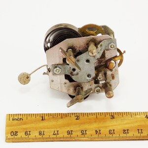 May include: Close-up of a vintage clockwork mechanism with a spring, gears, and a metal arm. The mechanism is about 5 cm wide and is made of metal.
