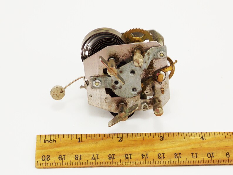 May include: Close-up of a vintage clockwork mechanism with a spring, gears, and a metal arm. The mechanism is about 5 cm wide and is made of metal.