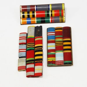 May include: Three rows of colourful military ribbons displayed on a black and gold metal bar. The ribbons are arranged in a vertical stack and feature a variety of colours, including red, white, blue, green, yellow, and black.