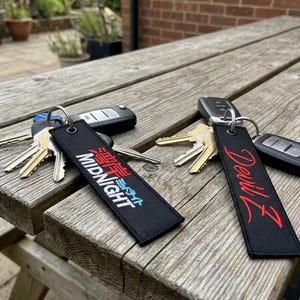May include: Two black rectangular fabric key tags with metal rings attached to car key fobs. One tag features the white text MIDNIGHT with red Japanese characters above it. The other tag displays the red cursive text Devil Z on the black surface.