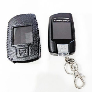 May include: A black leather key fob with a clear window and the words "Engine Start Stop" on the front. Next to it is a black remote control with a silver key ring and the words "Compustar" and "Engine Start Stop" on the front.