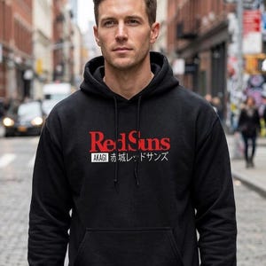 Pu&ograve; includere: Una felpa nera con cappuccio caratterizzata da una grafica frontale con la scritta rossa RedSuns sopra un riquadro bianco contenente il testo nero AKAGI e i caratteri giapponesi 赤城レッドサンズ. Il capo presenta un cappuccio con coulisse e una tasca a marsupio.