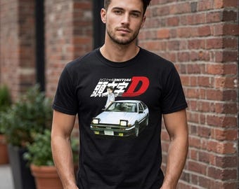 Może przedstawiać: Czarny t-shirt z krótkim rękawem z nadrukiem graficznym przedstawiającym biały samochód sportowy z otwieranymi reflektorami oraz postać z anime za nim. Nad samochodem znajduje się napis Initial D w języku angielskim i japońskim.
