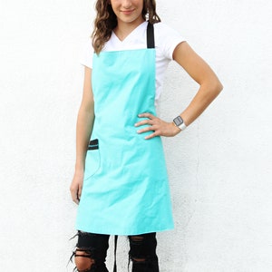 May include: A turquoise apron with black straps and a black pocket. The apron is worn by a person with long brown hair and a white shirt.