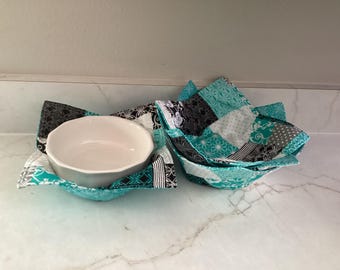 May include: Two handmade bowl cozies with a white ceramic bowl inside one. The cozies are made of teal, black, white, and gray patterned fabric. The bowl cozy is designed to hold a bowl of hot food and protect the user's hands.