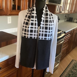 May include: A black and white checkered scarf with a solid black bottom section is draped over a white long-sleeved shirt. The scarf is displayed on a mannequin in a kitchen setting.