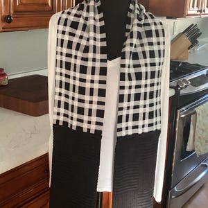 May include: A black and white checkered scarf with ribbed black ends is draped over a white long-sleeved shirt. The scarf is displayed on a black mannequin. The background shows a kitchen with wooden cabinets and appliances.