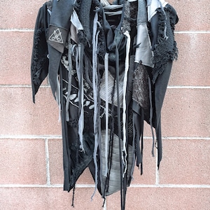 May include: A black and gray asymmetrical garment with frayed edges and long fabric strips hanging from the bottom. The garment is made of various fabrics and has a distressed look.