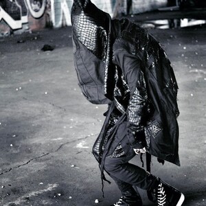 May include: A person wearing a black hooded jacket with a crocodile-embossed design. The jacket has a long, flowing silhouette and is paired with black pants and high-top sneakers.