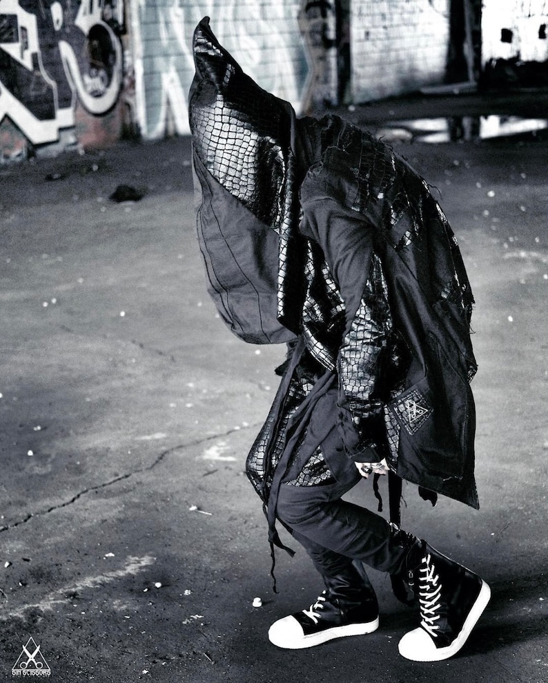 May include: A person wearing a black hooded jacket with a crocodile-embossed design. The jacket has a long, flowing silhouette and is paired with black pants and high-top sneakers.