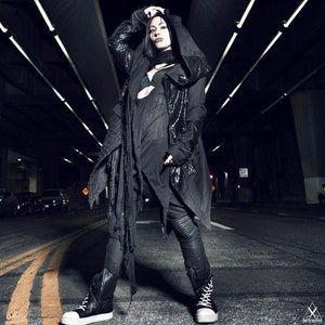 May include: A person wearing a black hooded jacket with a long, flowing scarf. The jacket has a distressed look and the person is standing in a dark, urban setting.
