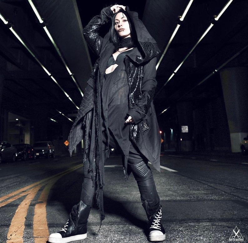 May include: A person wearing a black hooded jacket with a long, flowing scarf. The jacket has a distressed look and the person is standing in a dark, urban setting.