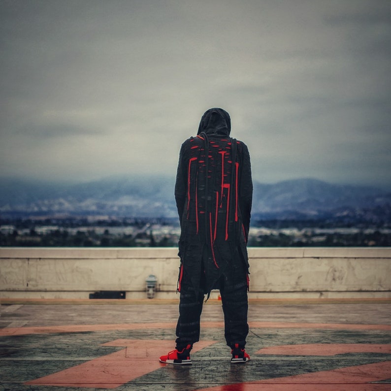 May include: A person wearing a black hooded jacket with red stitching and black pants. The person is standing on a rooftop with a city view in the background.