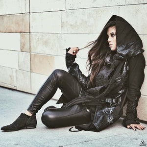 May include: A woman wearing a black leather jacket with a hood, black leather pants, and black boots. She is sitting on a white tiled floor with her legs crossed. Her hair is long and dark, and she is looking to the side.