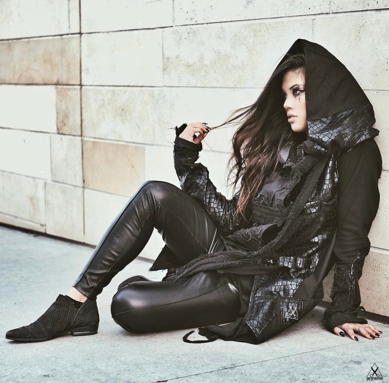 May include: A woman wearing a black leather jacket with a hood, black leather pants, and black boots. She is sitting on a white tiled floor with her legs crossed. Her hair is long and dark, and she is looking to the side.