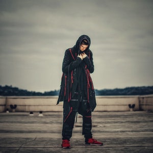 May include: A person wearing a black hooded jacket with red accents and black pants. The person is standing on a concrete surface.
