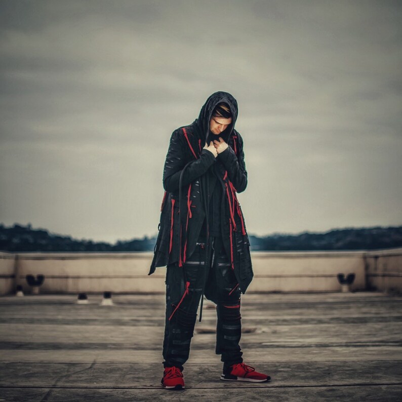 May include: A person wearing a black hooded jacket with red accents and black pants. The person is standing on a concrete surface.