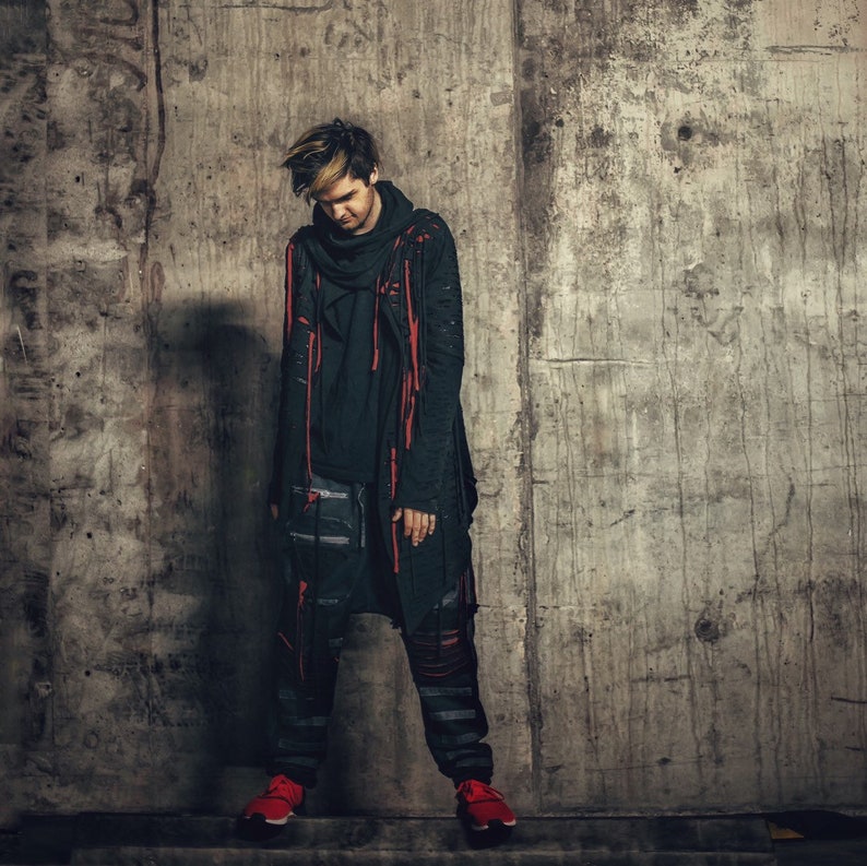 May include: A person wearing a black long-sleeved shirt with a red and black scarf, black pants, and red sneakers. The person is standing in front of a concrete wall.