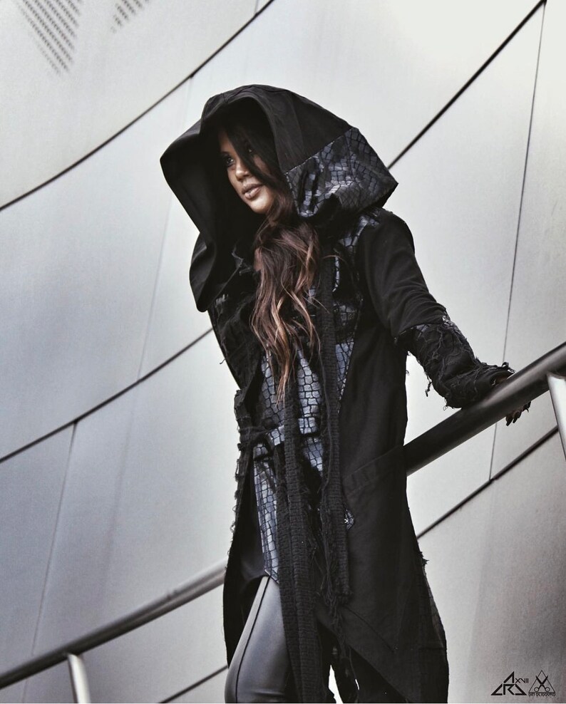 May include: A black hooded jacket with a long, flowing design and a textured pattern on the hood. The jacket has a frayed hem and sleeves, giving it a distressed look.