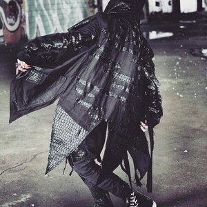 May include: A black hooded jacket with a long, flowing design. The jacket has a textured, crocodile-like pattern and frayed edges. The jacket is worn over black pants and black high-top sneakers.