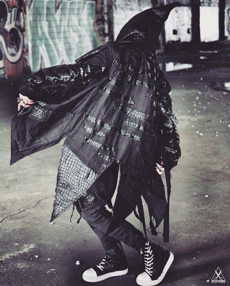 May include: A black hooded jacket with a long, flowing design. The jacket has a textured, crocodile-like pattern and frayed edges. The jacket is worn over black pants and black high-top sneakers.