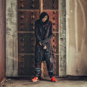 May include: A person wearing a black hooded jacket with red accents and black pants. The person is standing in front of a rusty metal door.